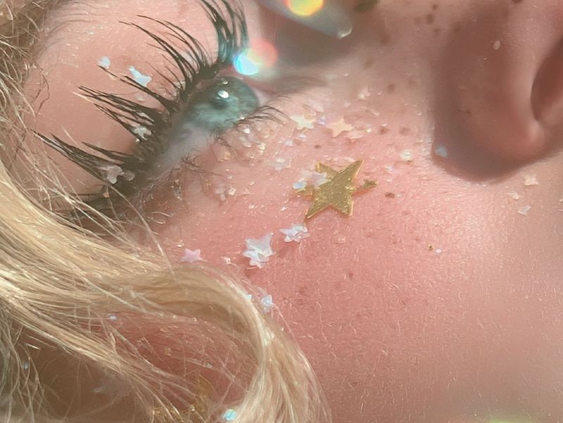 Close up detail of closed lashes, tiny stars and freckles on a cheek, and parted glossy pink lips.