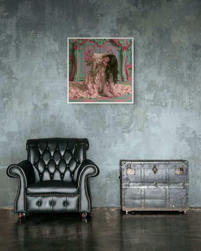 The melting pink dress print hung on a grey wall above a black tufted leather armchair and a metal trunk.