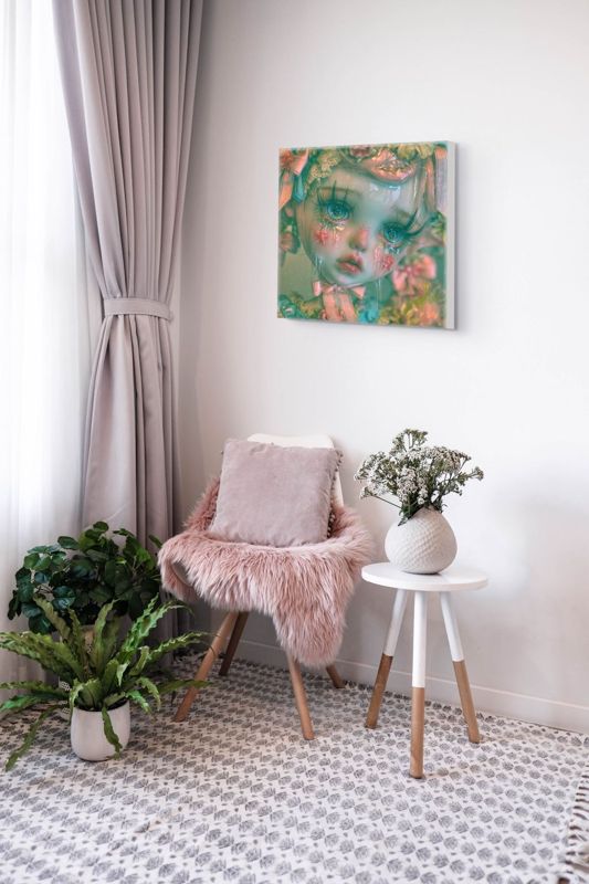 The crying doll print in teal and pink hung on a white wall above a blush velvet chair with a pink fur throw, a side table with flowers and potted ferns.