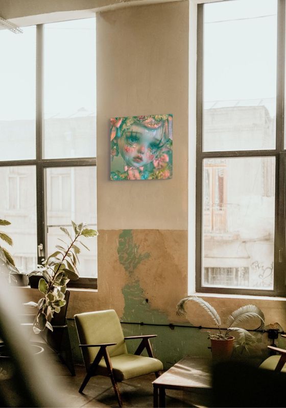 The crying doll print hung between tall industrial windows in a sunlit loft with a green mid century armchair and houseplants.