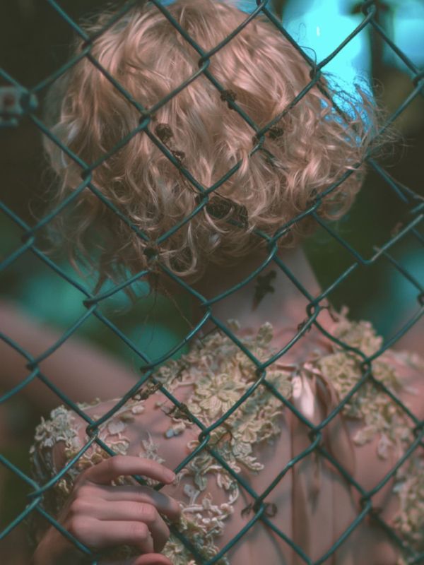 Detail of short blonde curls and a bare neck seen through teal chain link fence, blue garden light behind.