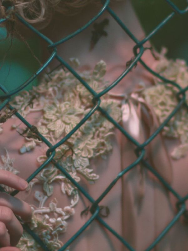 Detail of a sheer blush dress covered in pale lace flowers with a satin ribbon down the back, seen through chain link.