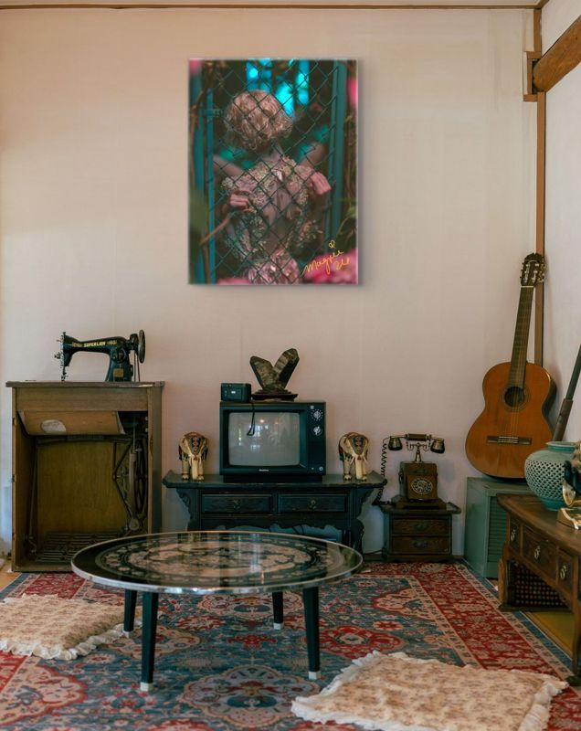 The teal and pink chain link fence print hung on a cream wall in a retro room with an antique sewing machine, a small vintage television, a guitar and a patterned red rug.