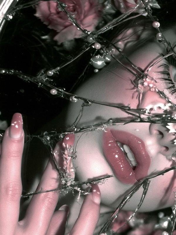 Detail of rose pink lips and long pink nails behind splintered mirror glass dotted with pearls, pink roses above.