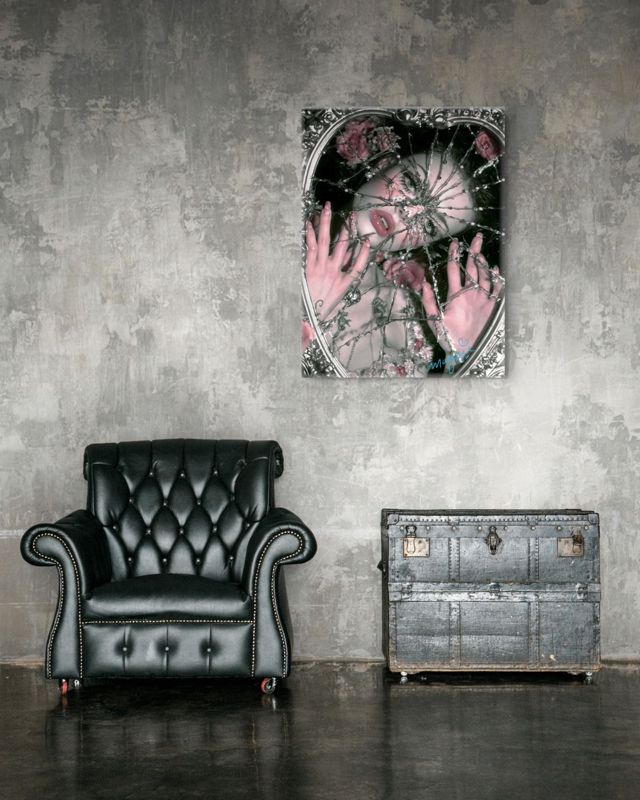 The shattered mirror print in black, silver and rose pink hung on a distressed grey plaster wall above a black tufted leather armchair and a metal trunk.