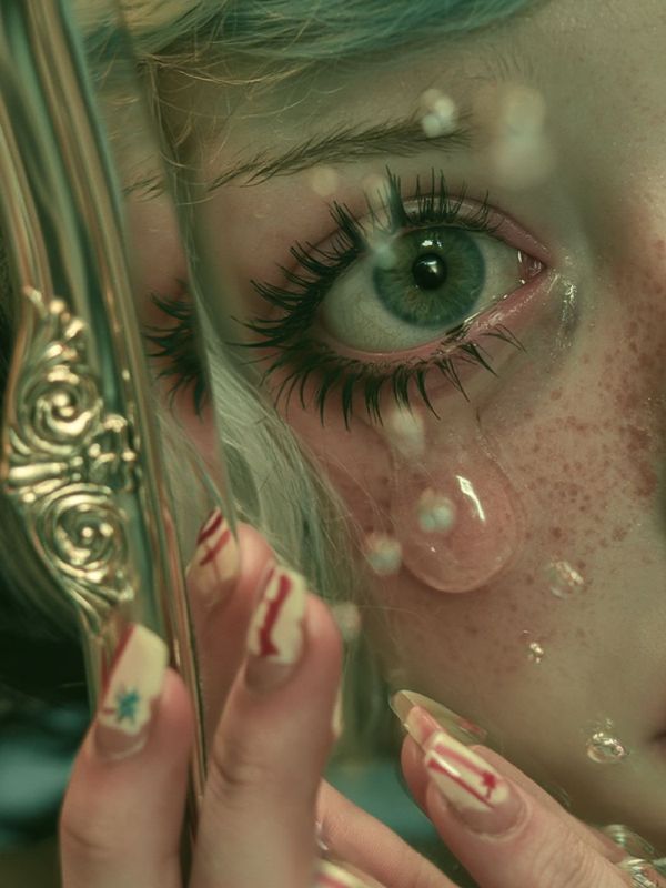 Detail of a green eye and a tear beside the ornate gold frame, fingers with tiny painted nails holding it, droplets on the skin.