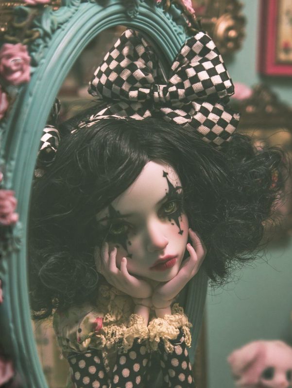 Closer view of the harlequin doll leaning on her hands in the teal rose trimmed mirror frame, checkered bow above her black curls.