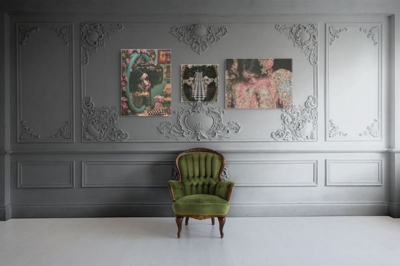 The harlequin doll print hung with a companion piece on a white paneled wall above a green velvet armchair.