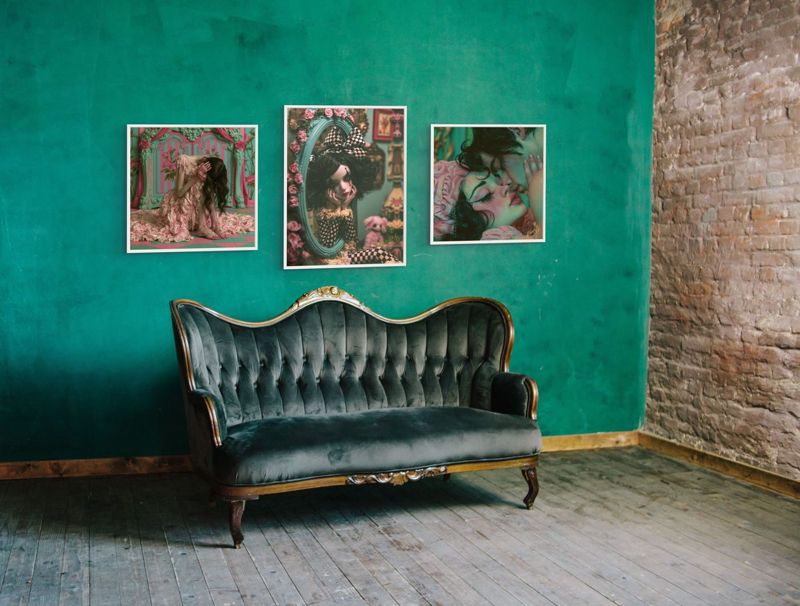 Three prints hung together in a white paneled room above a green velvet armchair: the harlequin doll, the melting pink dress and the lovers kiss.