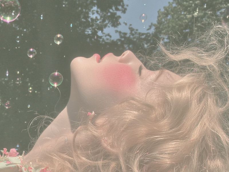 Close up detail of her upturned face and blonde curls against the pale sky, a daisy and soap bubbles beside her.