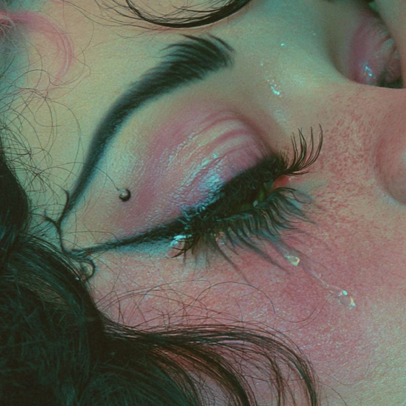 Close up detail of her closed eye with a sharp black wing, a small beauty mark and one tear on a flushed pink cheek.