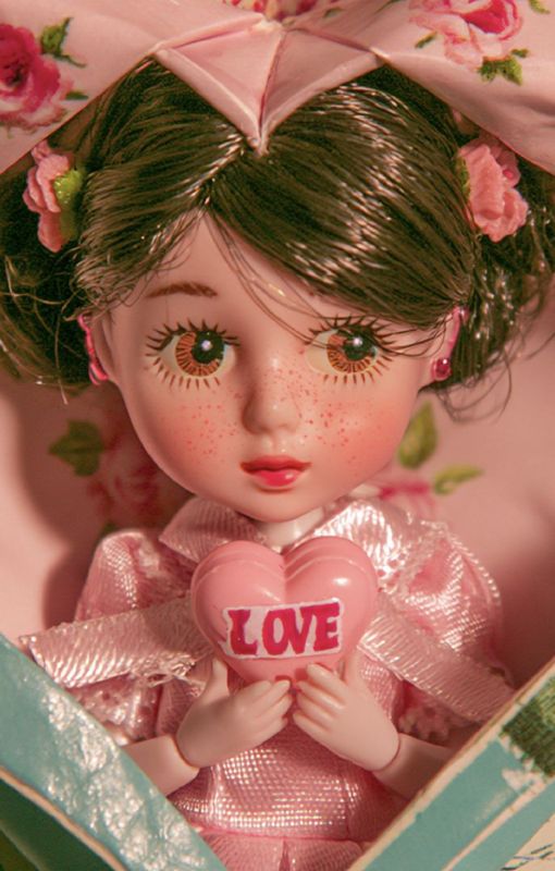 Close-up detail of a big-eyed doll face with amber eyes, painted lashes, freckles, dark brown hair with pink roses, pink lips, holding a pink LOVE heart, wearing a pink organza dress inside a floral-lined envelope.