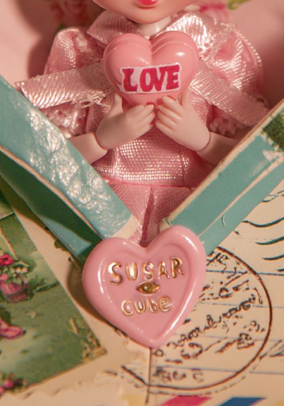 Close-up detail of small hands holding a pink heart stamped LOVE, pink organza dress, and a pink SUGAR CUBE heart charm dangling from the teal envelope edge, with vintage postcards beneath.