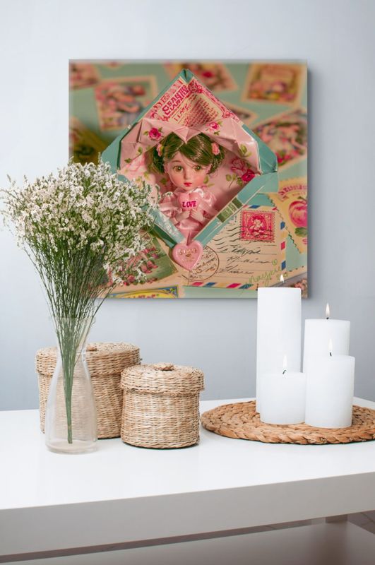 Square art print of a surreal doll in a vintage envelope on a gray-blue wall above a white surface with baby's breath in a glass vase, woven baskets, and white pillar candles on a woven mat.