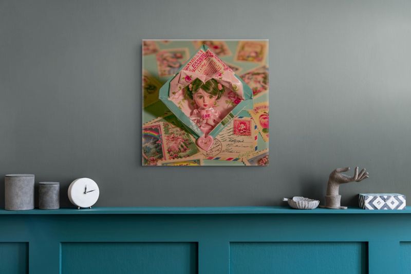 Square art print of a surreal doll in a vintage envelope on a dark teal wall above a teal shelf with a white clock, gray cylinders, a sculptural hand, and a geometric patterned box.