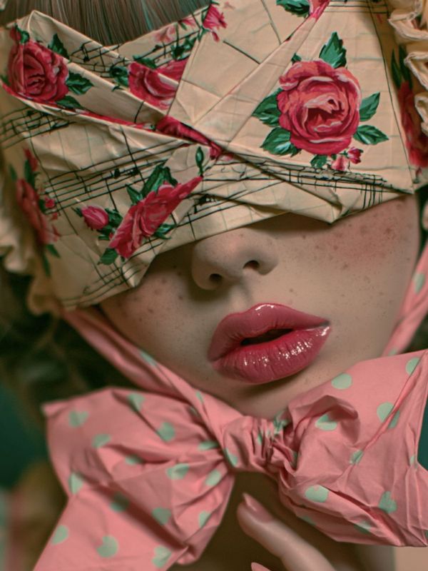 Close-up detail of an origami blindfold printed with pink roses and music notes covering the eyes, freckled skin, glossy pink lips, a pink polka-dot bow with teal dots below, teal-streaked hair peeking above.