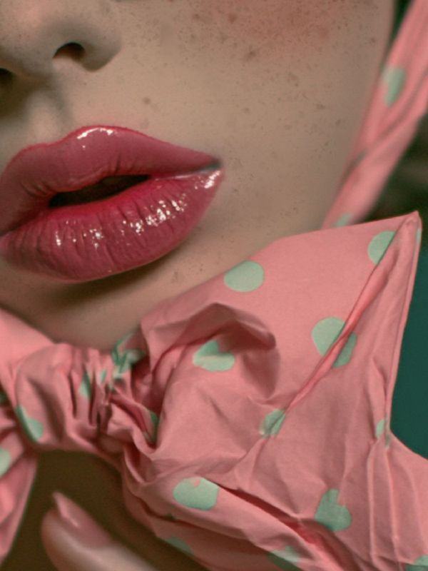 Close-up detail of glossy pink lips, freckled chin, and a crushed pink polka-dot bow with teal dots tied at the throat, teal background visible at the edges.