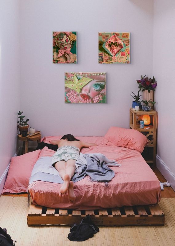 Three surreal art prints displayed as a gallery wall in a small bedroom with lavender walls: a portrait of a blindfolded figure with a pink bow (top left), the envelope doll (top right), and a green origami piece with a large blue eye and cherries (bottom center), all above a pallet bed with pink bedding, a wooden crate nightstand with plants and a salt lamp.
