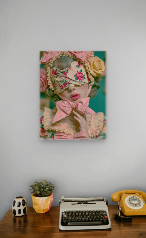 Portrait art print of a surreal blindfolded bow girl on a white wall above a mid-century wooden desk with a cream typewriter, yellow rotary phone, small potted plant, and a polka-dot ceramic piece.