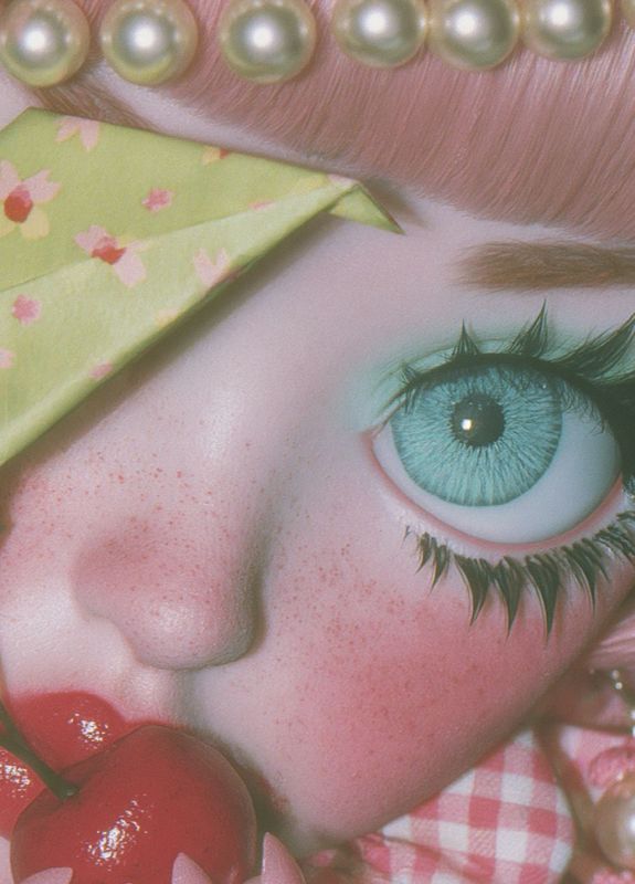 Close-up detail of one enormous teal eye with dramatic black lashes, freckled skin, a pearl headband above, a green floral origami star beside, and red cherries near glossy lips below.