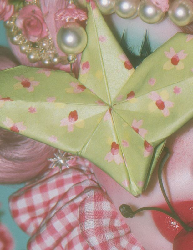 Close-up detail of a green origami star with pink and yellow floral print, surrounded by pink gingham ruffles, a silver starburst pin, pink curls, pearls, and a red cherry.