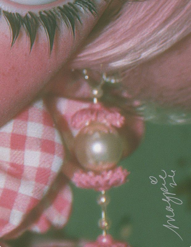 Close-up detail of a pearl drop earring with pink crocheted caps hanging beside dramatic black lashes, pink hair, and a pink gingham ruffle, with the Magpie signature in white script against a teal background.