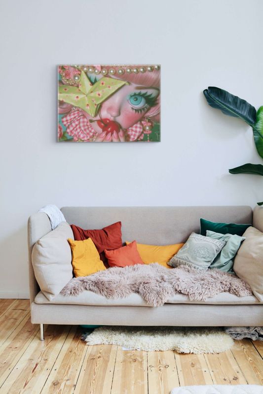 Landscape art print of a surreal big-eyed cherry girl on a white wall above a beige linen sofa with rust, orange, sage, and teal throw pillows, a pink faux fur throw, and a large tropical plant.