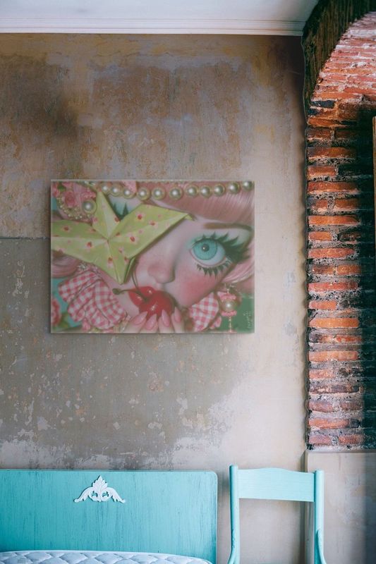 Landscape art print of a surreal big-eyed cherry girl on a weathered plaster and exposed brick wall above a turquoise painted bench and turquoise chair.