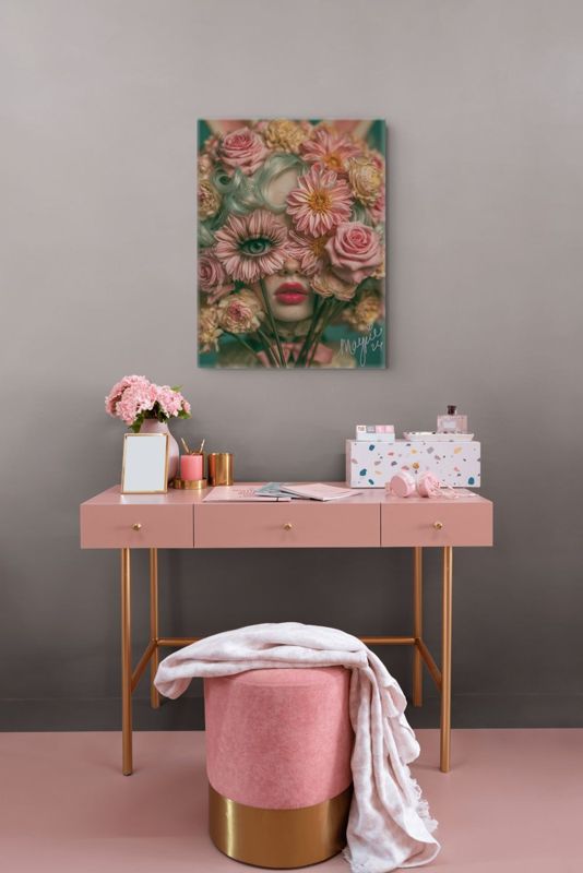 Portrait art print of a surreal floral cyclops girl displayed above a pink desk with gold legs, a pink velvet ottoman, pink candle, gold accessories, and a terrazzo storage box against a mauve wall.