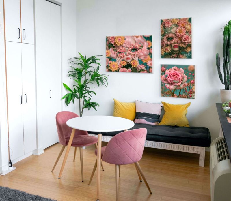 Three surreal floral art prints displayed as a gallery wall in a bright studio apartment: a landscape butterfly floral mask portrait, a square floral cyclops portrait, and a square bandaged pink rose, all in teal and pink tones, above a black daybed with yellow and pink pillows, beside pink velvet dining chairs, a white round table, and a tall green plant.
