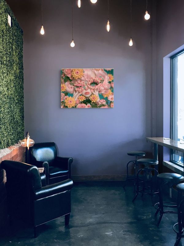 Landscape art print of a surreal butterfly floral mask portrait on a lavender wall in an industrial lounge with hanging Edison bulbs, a living green wall, black leather club chair, and bar stools by a window.