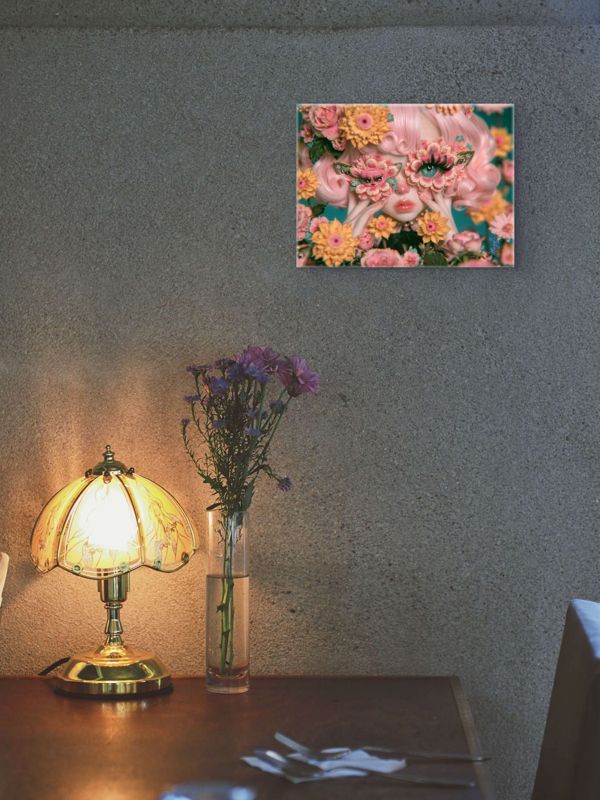 Landscape art print of a surreal butterfly floral mask portrait on a textured concrete wall above a desk with an art nouveau amber glass lamp and purple wildflowers in a clear vase.
