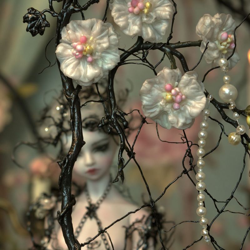 Closer view of white silk flowers and pearl strands on twisted black wire branches, with the pale doll blurred in candlelight behind.