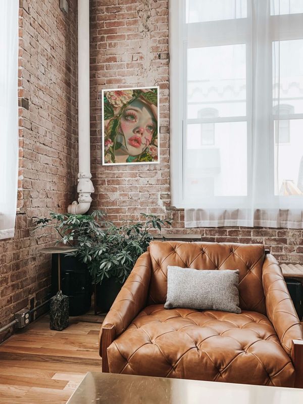 The girl in the vines print hung on a white wall above a tan leather sofa in a brick loft with plants.