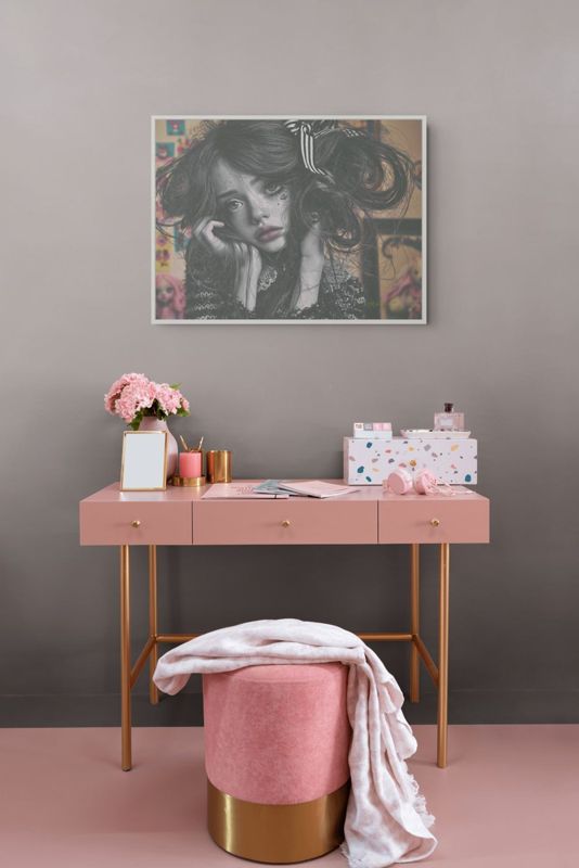 The black and white girl print hung on a mauve wall above a pink desk with gold legs, a pink stool and a vase of pink flowers.