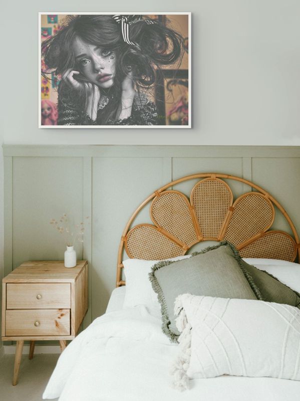 The black and white girl print hung on a sage green wall above a bed with a rattan flower headboard and white linen.