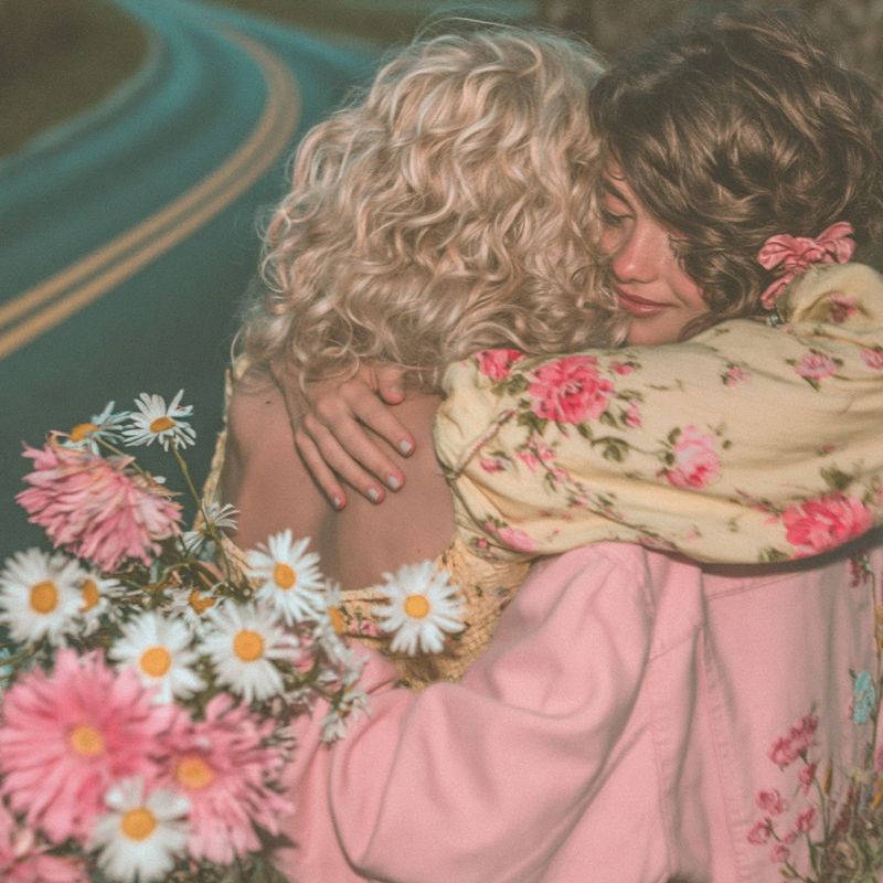 Closer view of the two girls embracing, blonde and brown curls pressed together, pink jacket and yellow floral sleeve, daisies between them.
