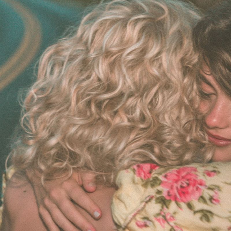 Close up detail of the two heads resting together, a pink bow in brown curls and an arm in a yellow floral sleeve wrapped around a shoulder.