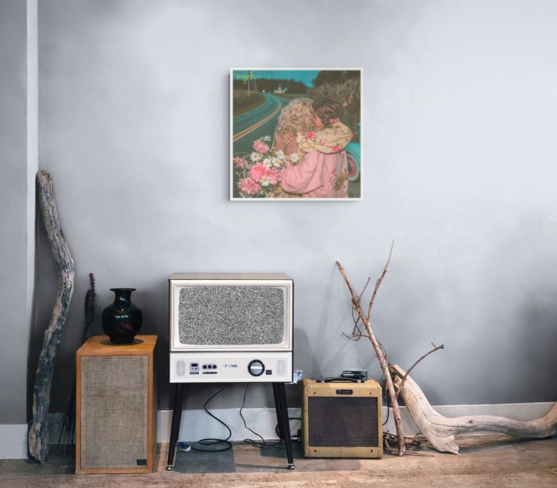 The roadside hug print hung on a grey wall above a vintage radio, a speaker and driftwood.