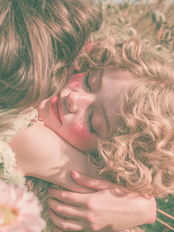Closer view of the embrace: a freckled flushed face with closed eyes and pale curls pressed against her friend's brown hair.