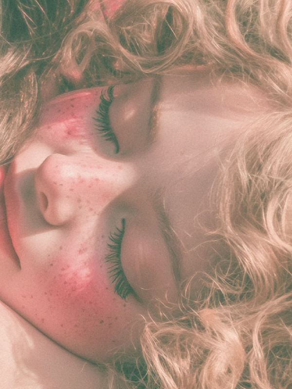 Close up detail of a peaceful freckled face with closed eyes and rosy cheeks, framed by loose pale curls.