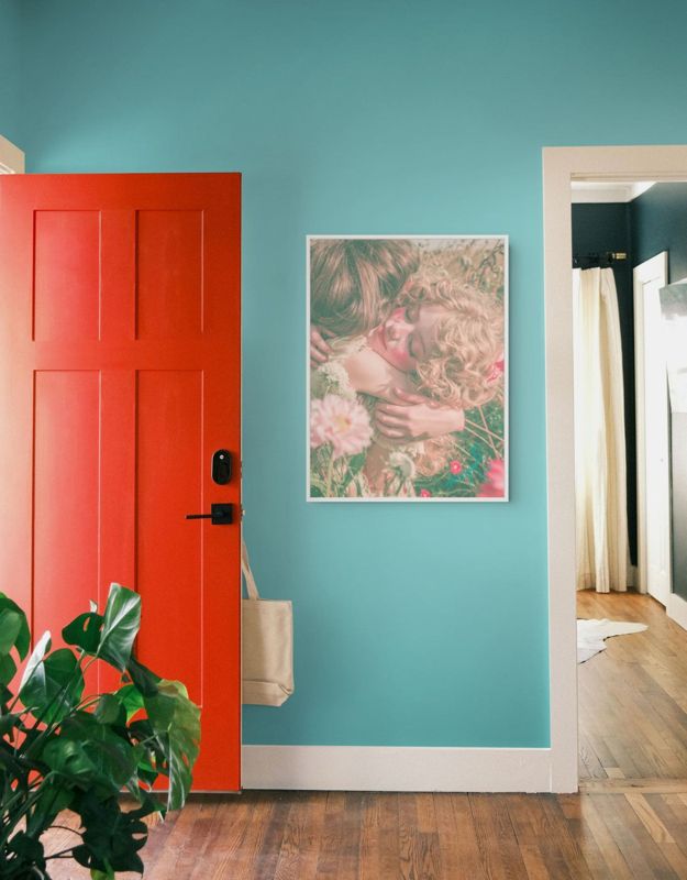 The friends in the flower field print hung on a teal wall beside a red door and a large leafy plant.