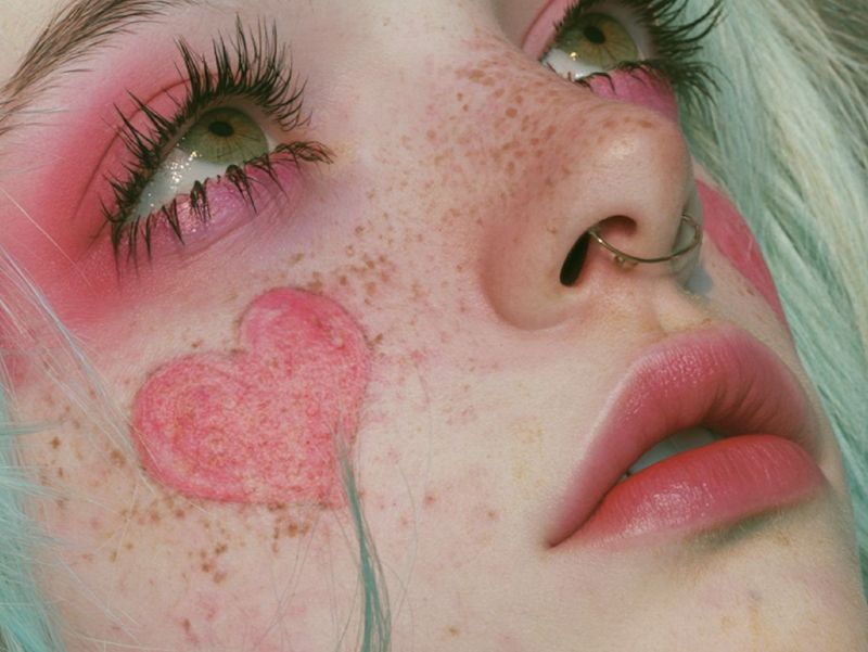 Close up detail of a green eye ringed in hot pink shadow above a pink heart shape on a freckled cheek.