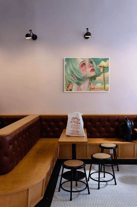 The mint hair girl print hung on a pale wall in a cafe corner with wooden chairs and a dark leather bench.